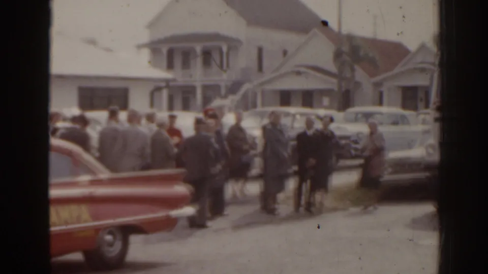 Grey-uniformed adults stand in a Florida street scene with a red lettered car and mid-century parked sedans.