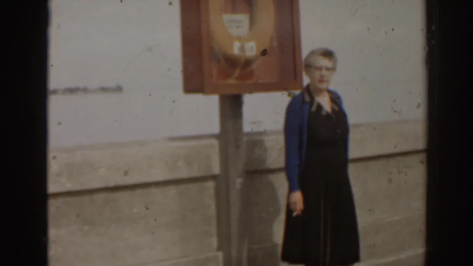 In 1963 Florida home-movie footage, a woman in a dark dress and blue cardigan stands near a life preserver cabinet on a concrete pier.