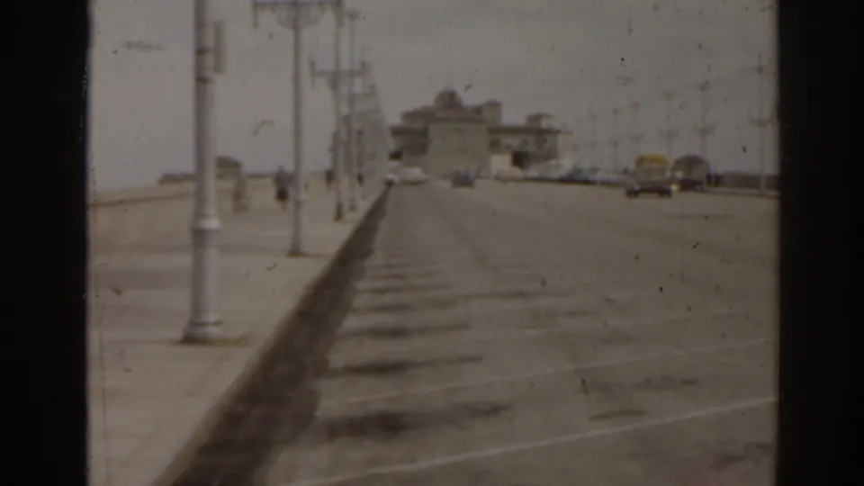 A waterfront road or boardwalk in Florida shows lampposts, vintage cars, a low seawall, and distant seaside buildings.