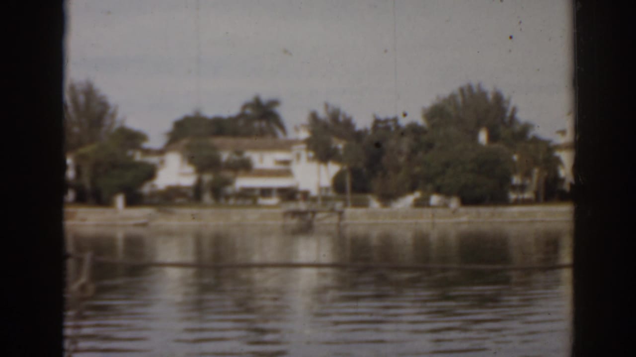 Lake water with distant houses, trees, and a dock post in front.