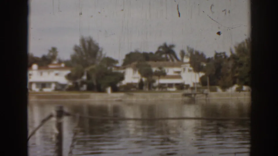 1963 Florida home-movie frame with lake reflections, distant residences, trees, and nearby dock or railing elements.