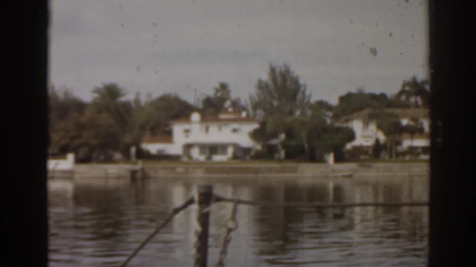 1963 Florida home-movie view of waterfront houses, trees, dock posts, and calm water seen through dark foreground framing.