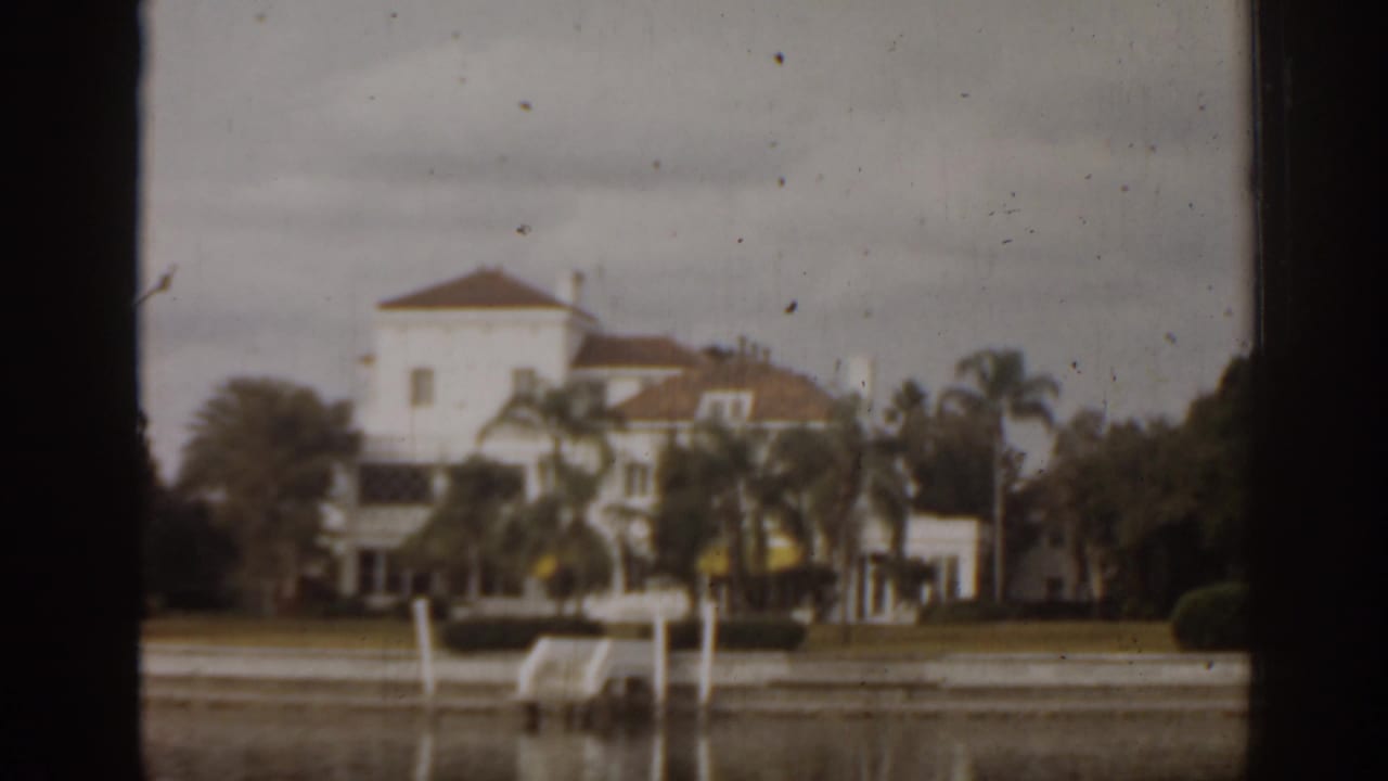 Large white waterfront mansion with palms and dock posts across the water.