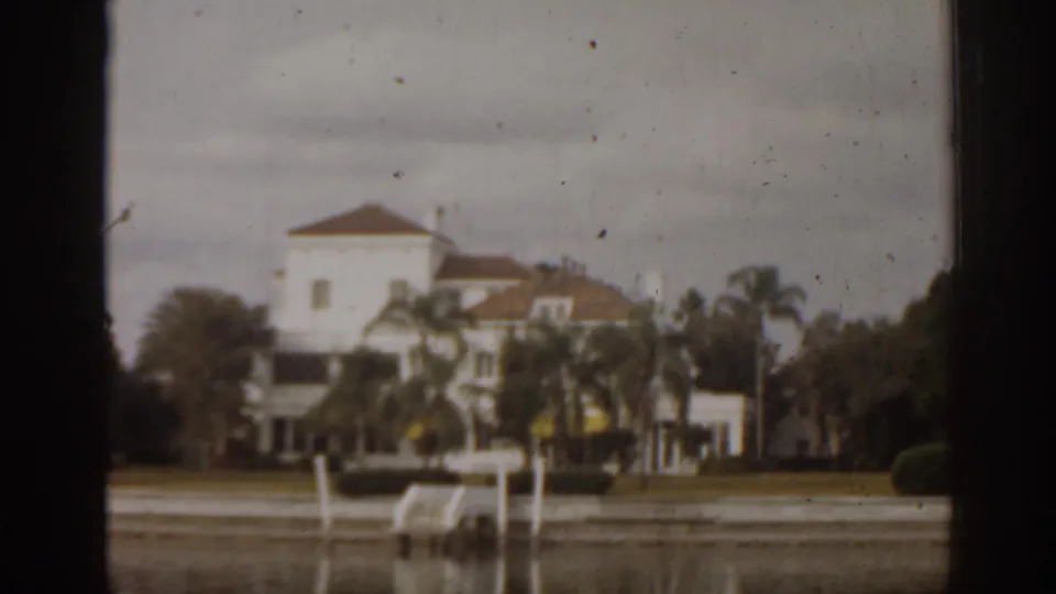 1963 Florida view of a large white waterfront mansion, palm trees, seawall, dock posts, and muted sky across calm water.