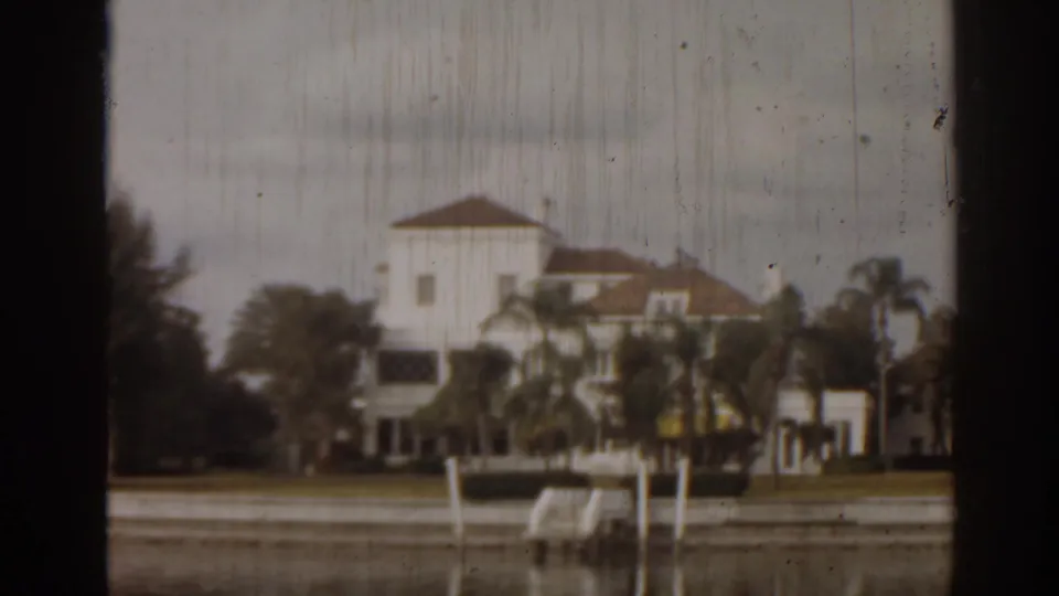 1963 Florida home-movie frame showing a white waterfront mansion, palms, lawn, seawall, dock posts, and visible 8mm wear.