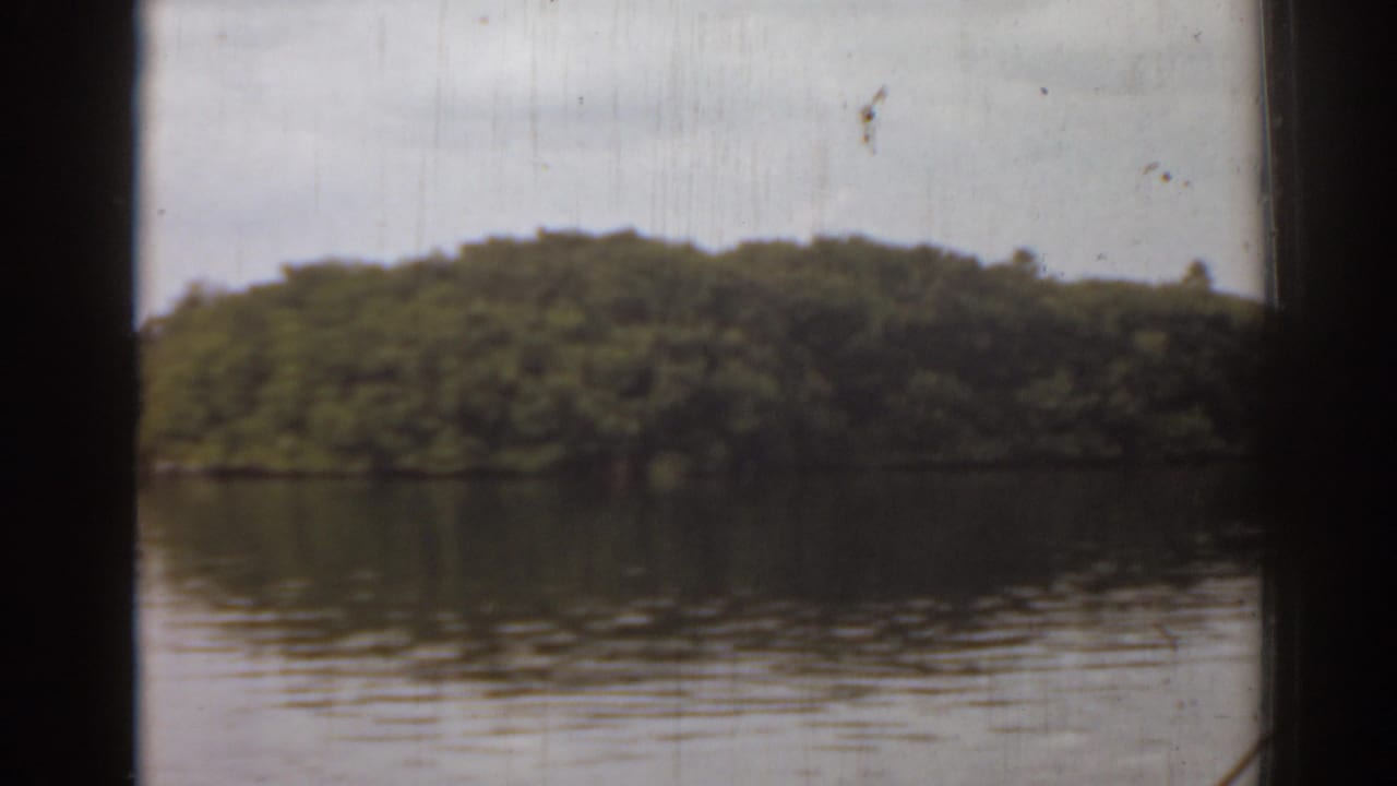 Boat-view frame from 1963 Florida footage, with a wooded island across reflective water and dark structural framing in the foreground.