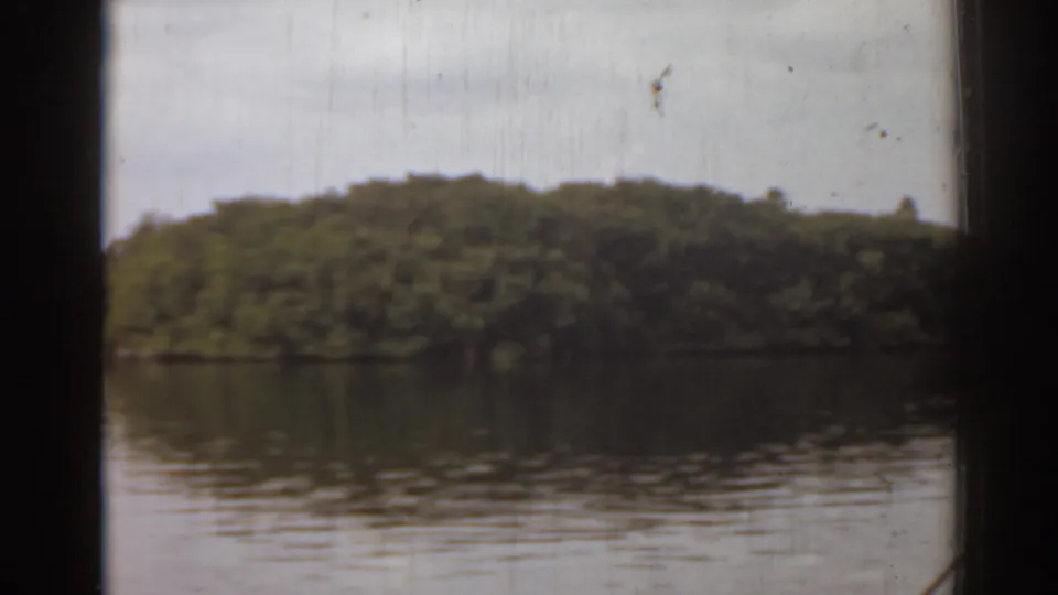 Boat-view frame from 1963 Florida footage, with a wooded island across reflective water and dark structural framing in the foreground.