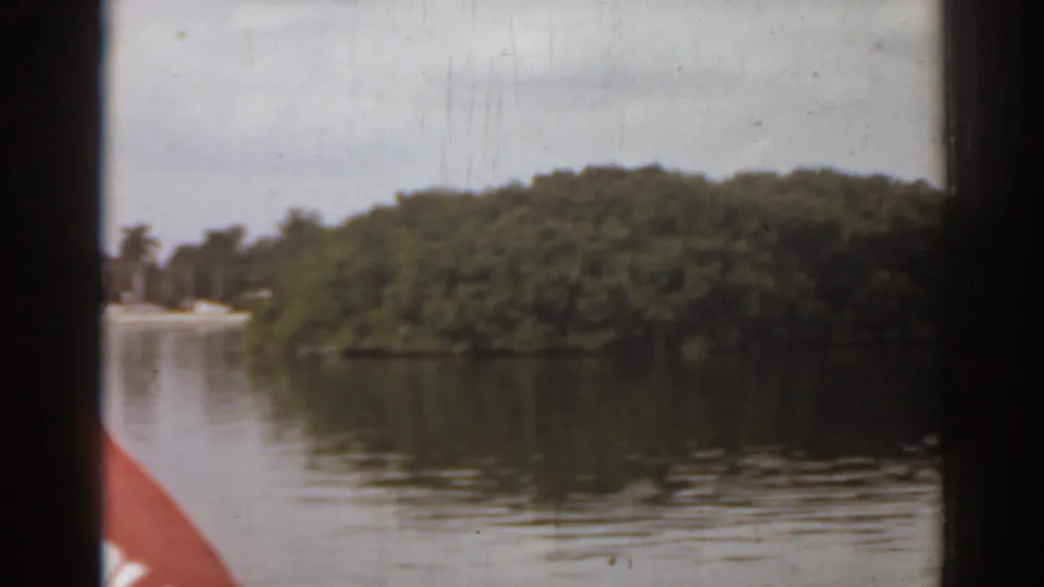 1963 Florida home-movie frame showing a low green island, rippled water, and a dark boat-window edge with faint shoreline at left.
