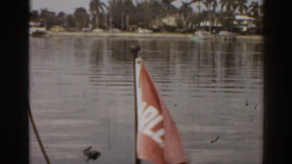 1963 Florida boat-view frame with a small red flag in front, calm water, palm trees, and distant waterfront houses.