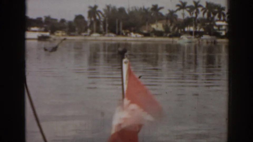 1963 Florida 8mm frame from a boat showing a small red flag, calm water, palms, and distant waterfront homes.