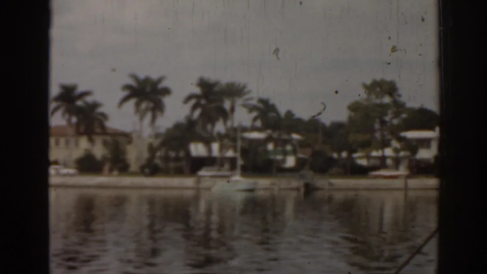 1963 Florida home-movie frame showing calm water, palm trees, waterfront houses, docks, and a small boat along the opposite shore.