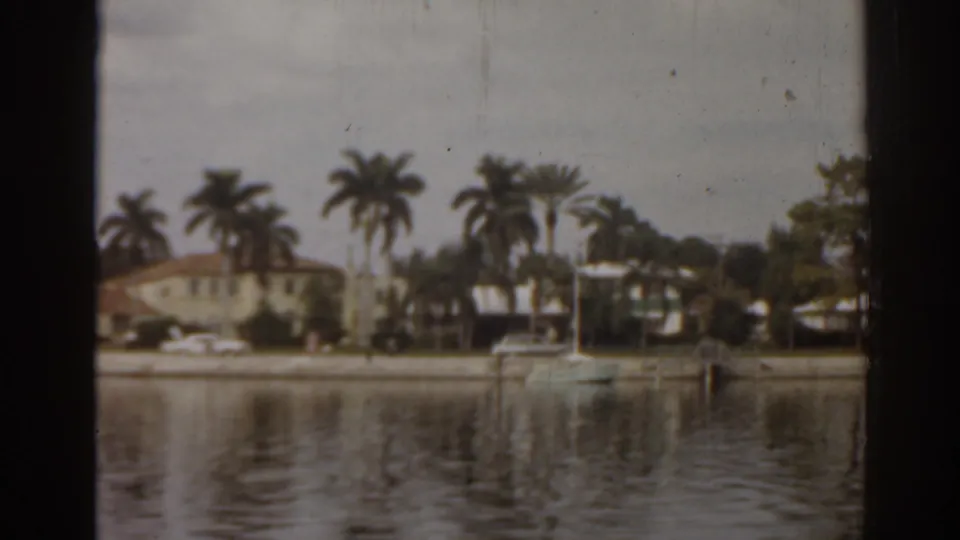 1963 Florida shoreline view with low homes, tall palms, a docked boat, parked cars, and soft reflections on the water.