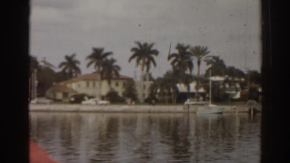 1963 Florida home-movie frame showing a quiet waterfront street, palm trees, houses, parked cars, and a small moored sailboat.