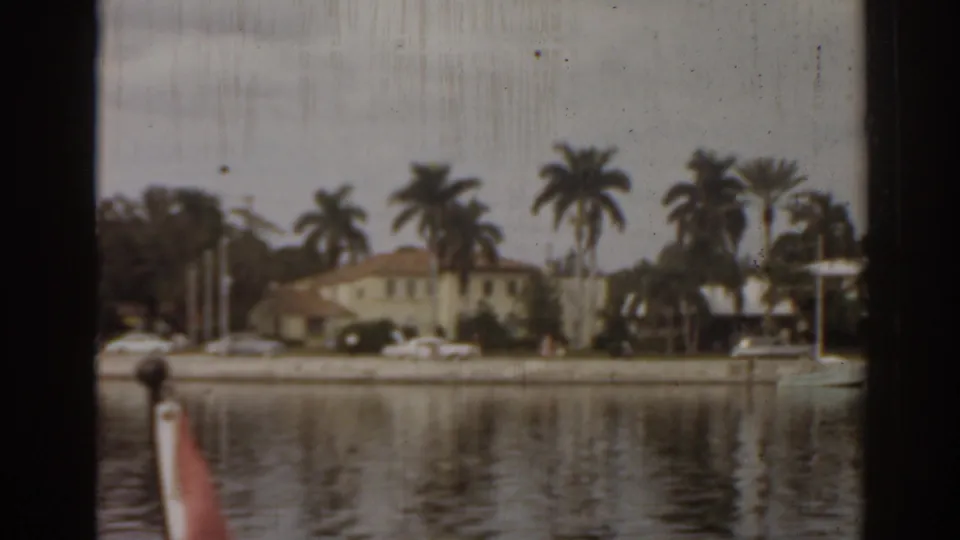 1963 Florida shoreline frame with residential buildings, tall palms, parked cars, a docked boat, and muted reflections in the water.