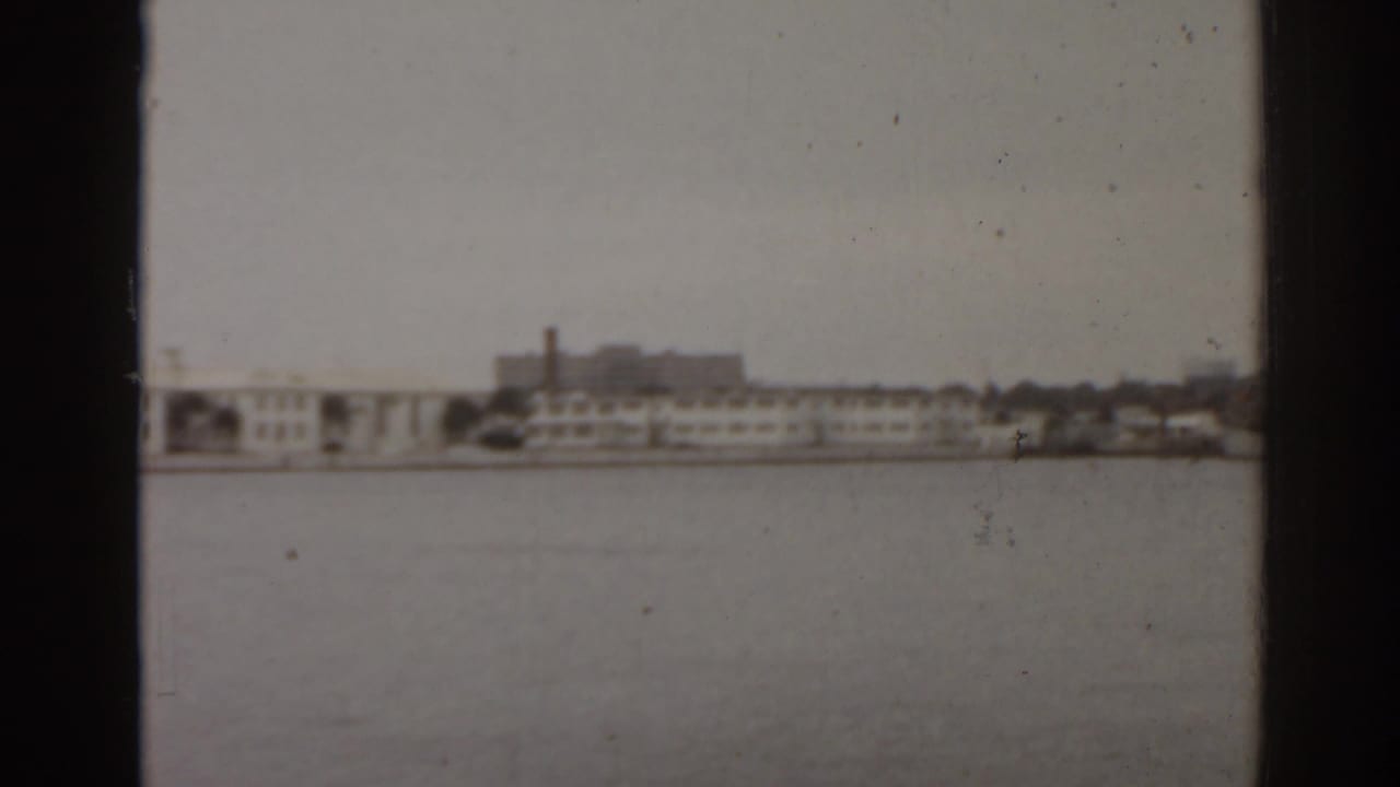 1963 Florida home-movie frame showing open water and a distant low-rise shoreline with a prominent smokestack.