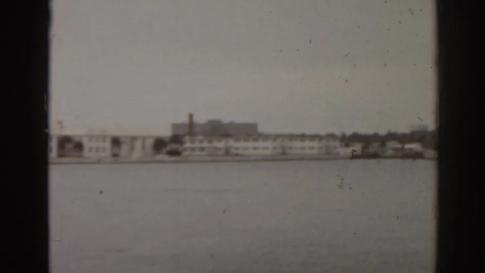 1963 Florida home-movie frame showing open water and a distant low-rise shoreline with a prominent smokestack.