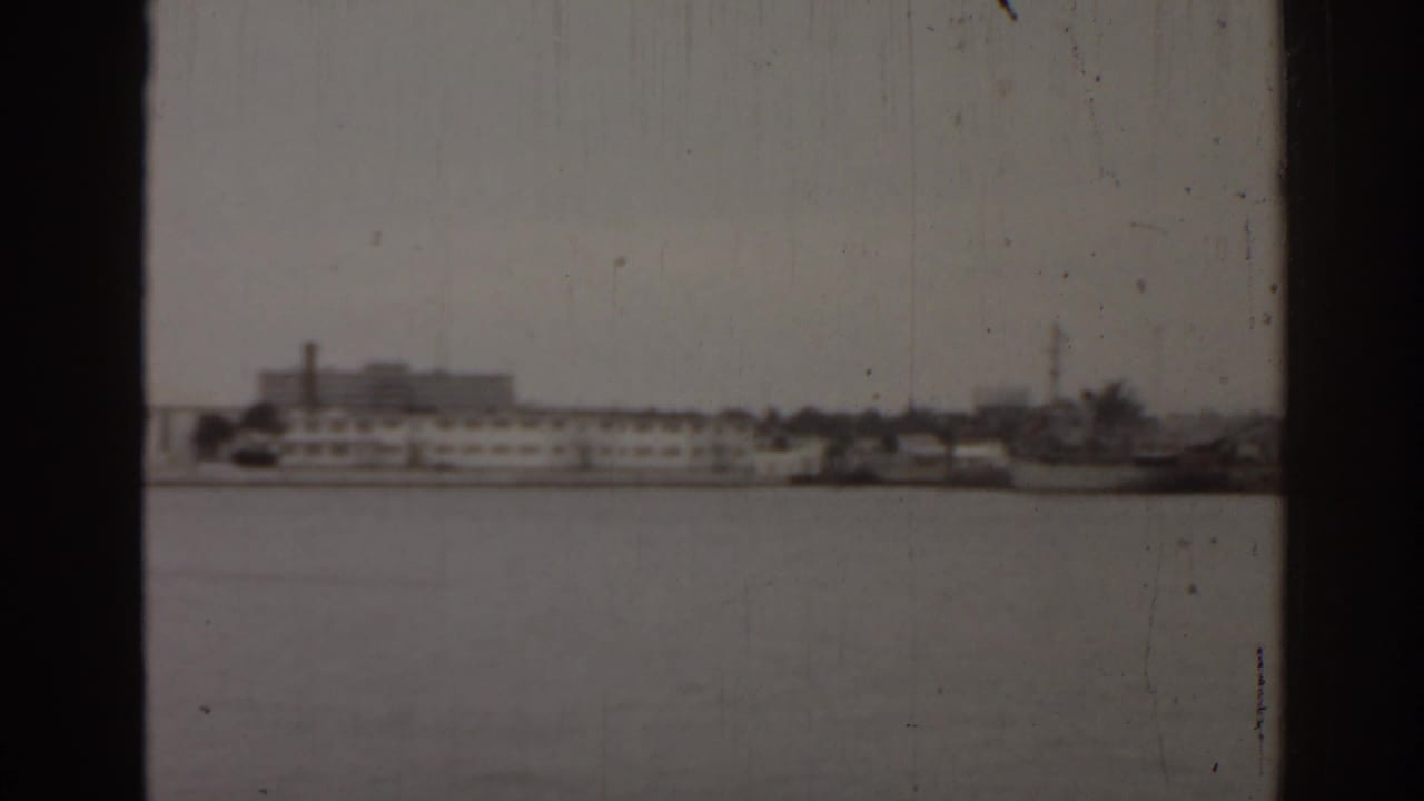 Distant Florida waterfront in a 1963 8mm frame, with low buildings, boats, and pale sky across open water.