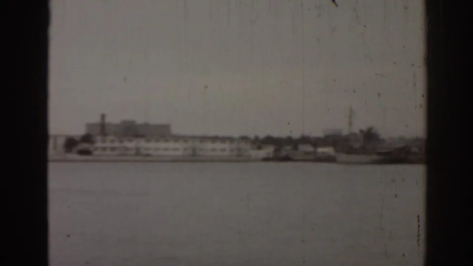 Distant Florida waterfront in a 1963 8mm frame, with low buildings, boats, and pale sky across open water.