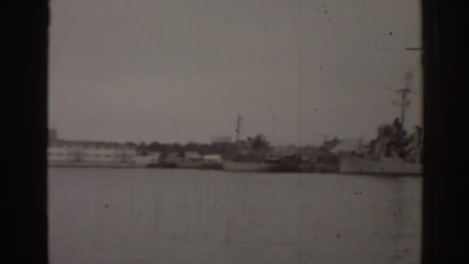 1963 Florida harbor view with a naval-looking vessel, small boats, and low shoreline structures across the water.