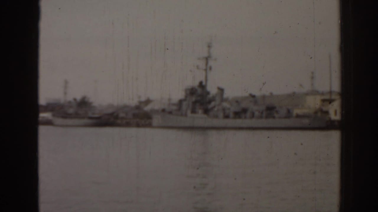 Large naval ship moored across calm harbor water with dock structures behind it.