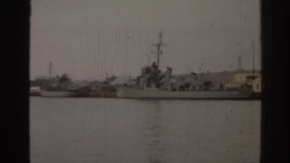 1963 Florida 8mm frame showing a large naval ship docked across calm water, with shoreline buildings and trees in soft focus.