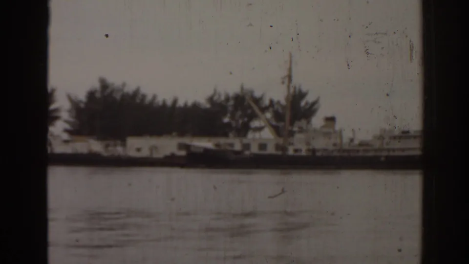 1963 Florida harbor view with a small boat near dockside buildings and trees, framed by dark 8mm film borders.