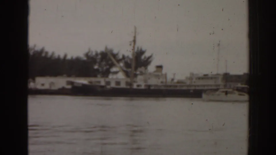 1963 Florida 8mm still showing a docked workboat near low waterfront buildings, trees, and calm harbor water.