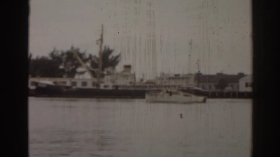 1963 Florida harbor frame with a small sailboat crossing foreground water near docked boats and low shoreline buildings.