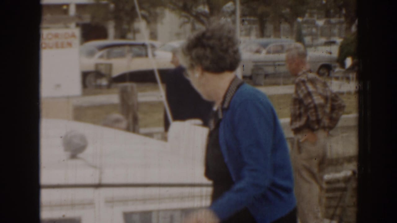 A woman in a blue cardigan walks beside a white boat at a Florida waterfront, with mid-century parked cars and another pedestrian behind her.