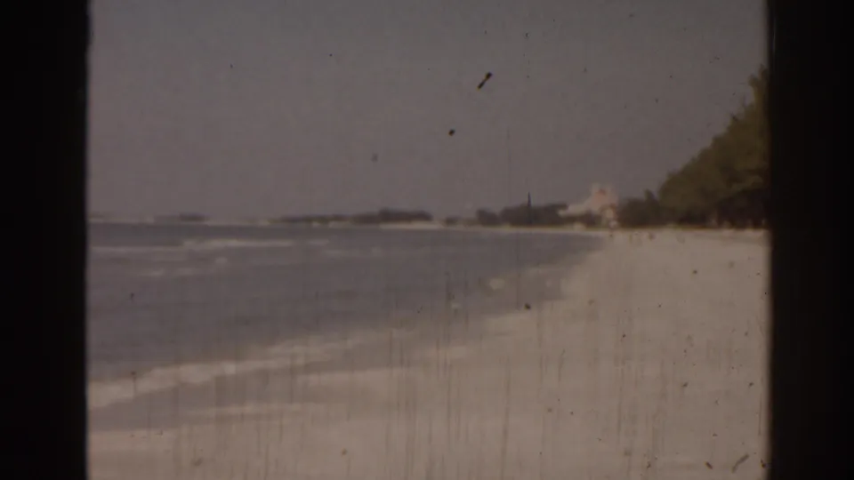 A wide 1963 Florida beach view shows calm water, pale sand, shoreline trees, distant structures, and aged 8mm film texture.