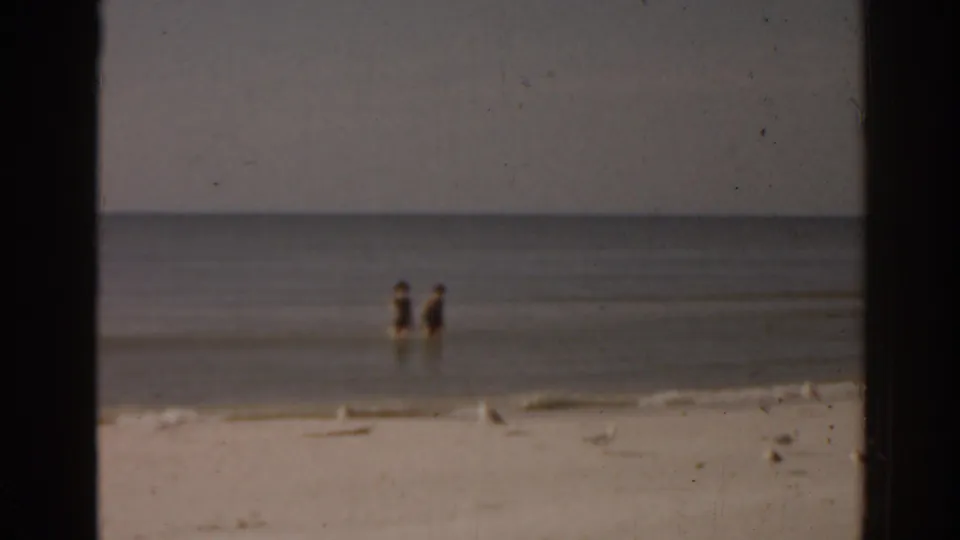 Soft 1963 Florida 8mm frame showing two distant people in shallow water, low waves, pale sand, and seagulls near the shoreline.