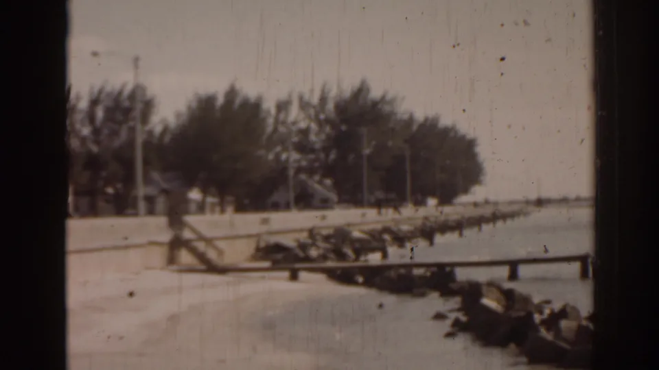 1963 Florida 8mm frame showing a seawall, shoreline rocks, calm water, trees, poles, and dark film-frame borders.