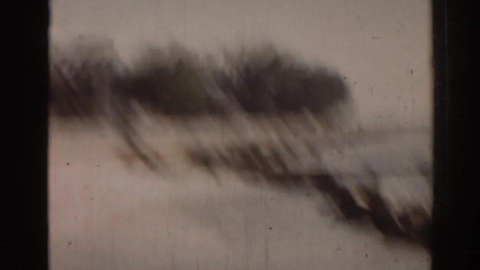 Soft, blurred 1963 Florida coastal frame with shoreline rocks, water, dark trees, and visible 8mm film wear.