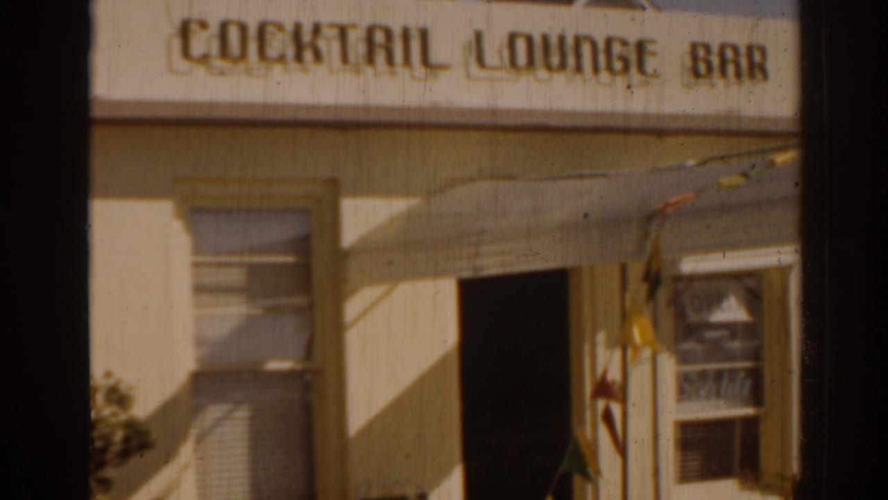 Sunlit cocktail lounge bar facade with sign, awning, doorway, window, and partial patio decor.