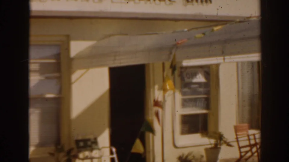 1963 Florida home-movie frame of a lounge entrance with awning, window blinds, colorful pennants, an open doorway, and patio seating details.
