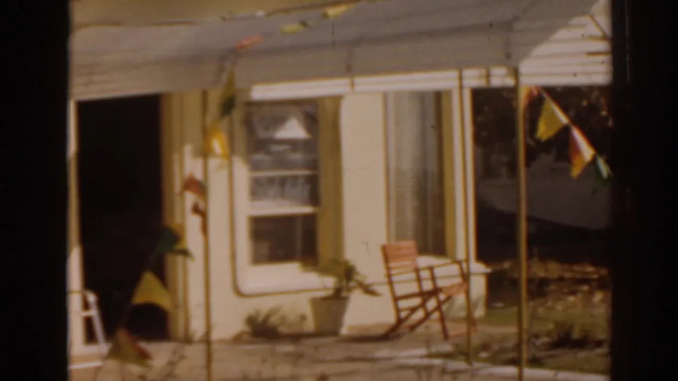 1963 Florida 8mm frame showing the lower lounge facade, including an open door, window, patio chair, plants, and colorful pennants.