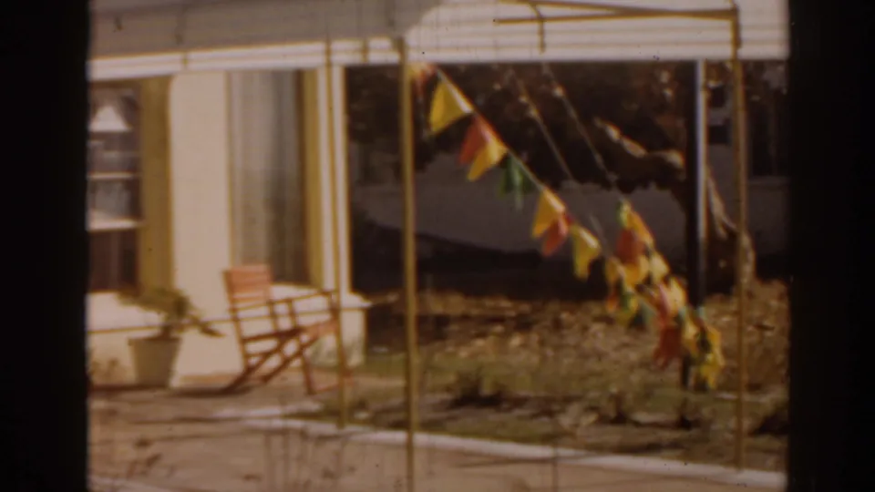 1963 Florida home-movie frame of the lounge exterior with window blinds, patio chair, potted plant, awning, and pennant bunting in the foreground.