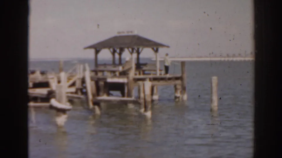1963 Florida 8mm frame showing a roofed wooden dock, pilings, calm water, and a hazy distant shoreline.