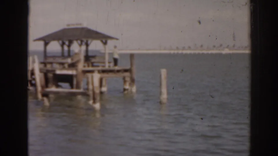 1963 Florida waterfront frame with a distant person beside a roofed wooden dock and open water beyond.