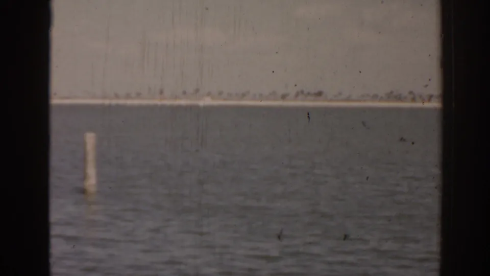 1963 Florida waterfront frame showing open water, one dock piling, and a hazy low shoreline across the channel.