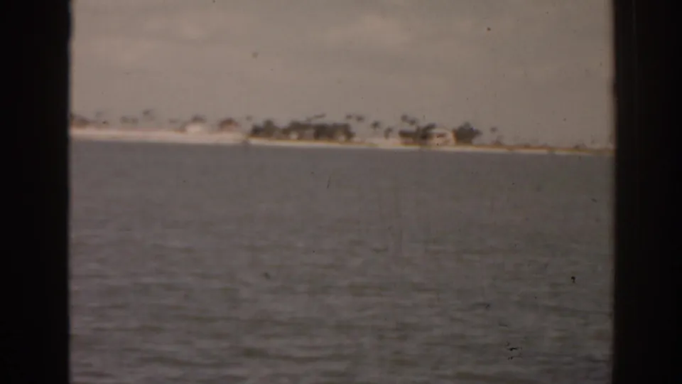 1963 Florida waterfront frame showing calm water, a distant shore, small buildings, and palm-like trees.