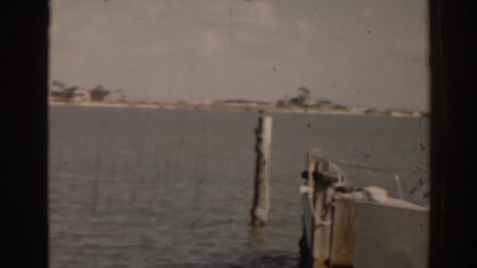 1963 Florida home-movie frame showing calm coastal water, a dock piling, a moored boat bow, and a low distant shoreline.