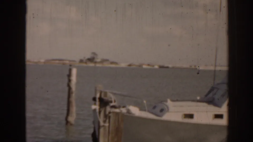 1963 Florida coastal frame with a white boat moored near wooden pilings and a distant low shoreline across the water.