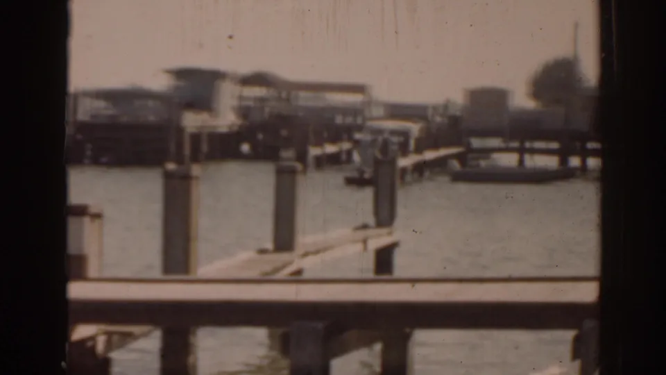 1963 Florida home-movie view of a waterfront pier area with railings, pilings, moored boats, and small shorefront buildings.