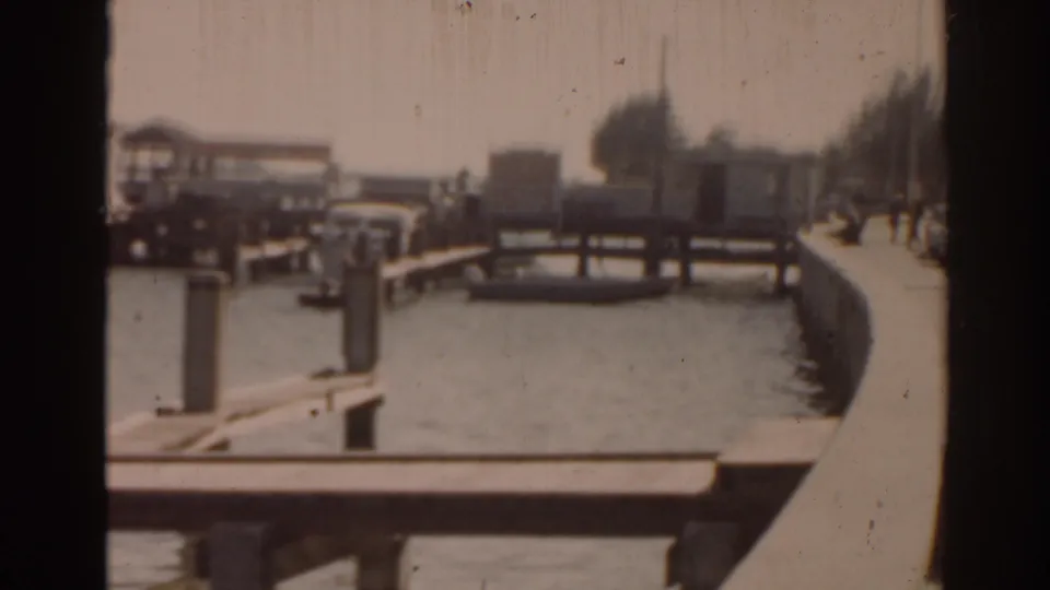 1963 Florida waterfront frame showing dock railings, moored boats, shorefront buildings, trees, and tiny distant people along the right edge.