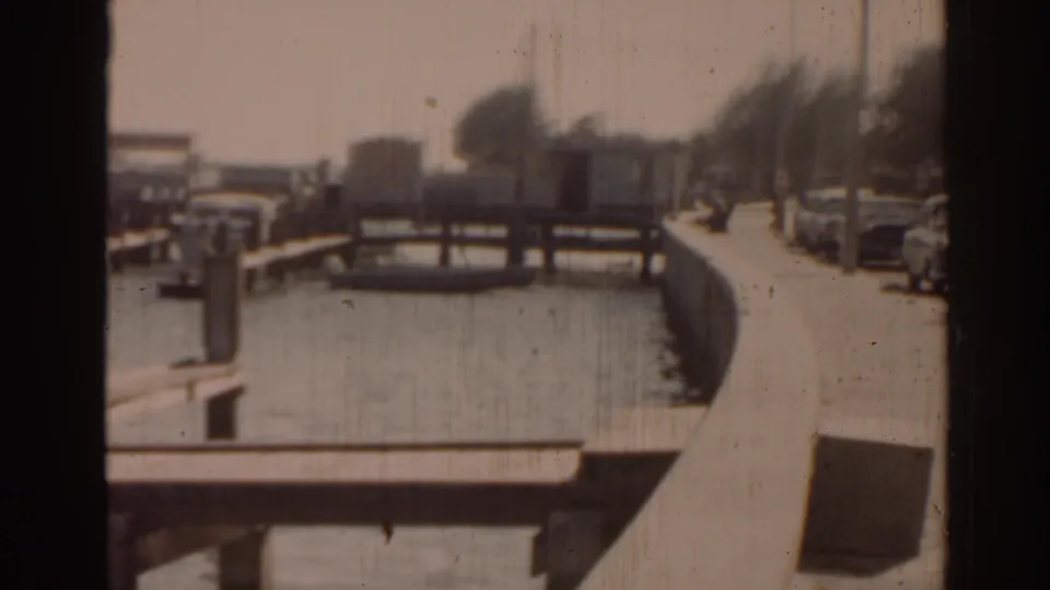 1963 Florida home-movie frame showing a canal, wooden docks, parked cars, and a bright waterfront street.