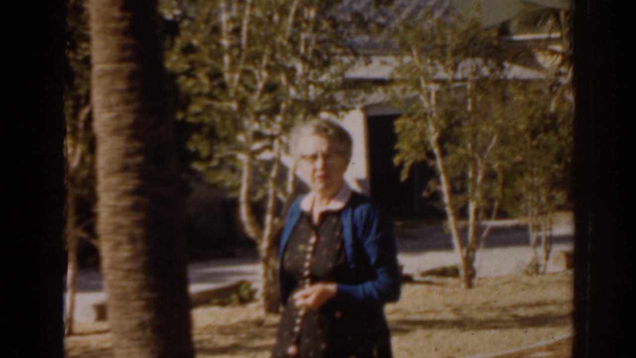Elderly woman standing near a palm tree in a sunny yard.