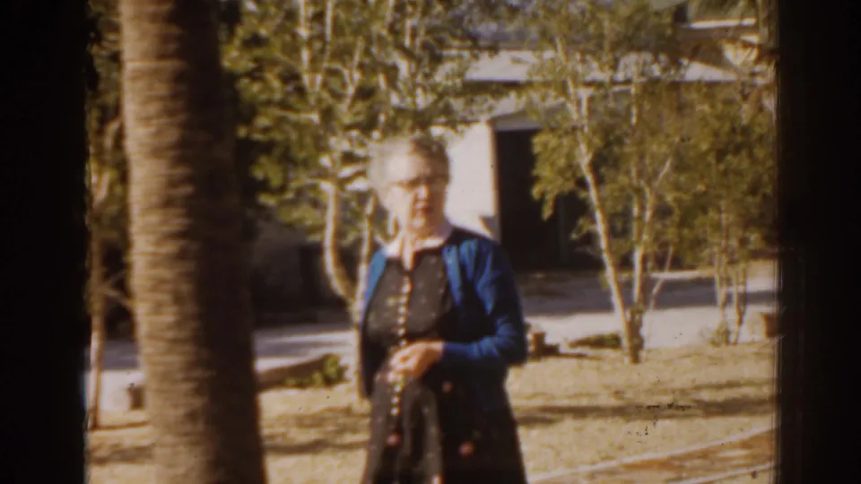 1963 Florida frame showing an elderly woman walking through a sunny yard with palms and a house behind her.