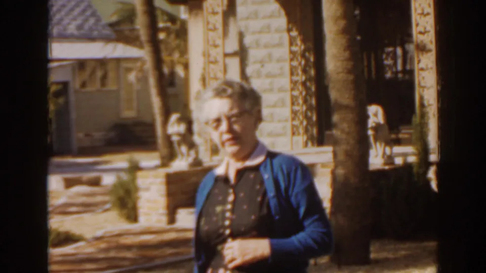 1963 Florida home-movie frame of an elderly woman near a porch with stone pillars, palms, and small statues.