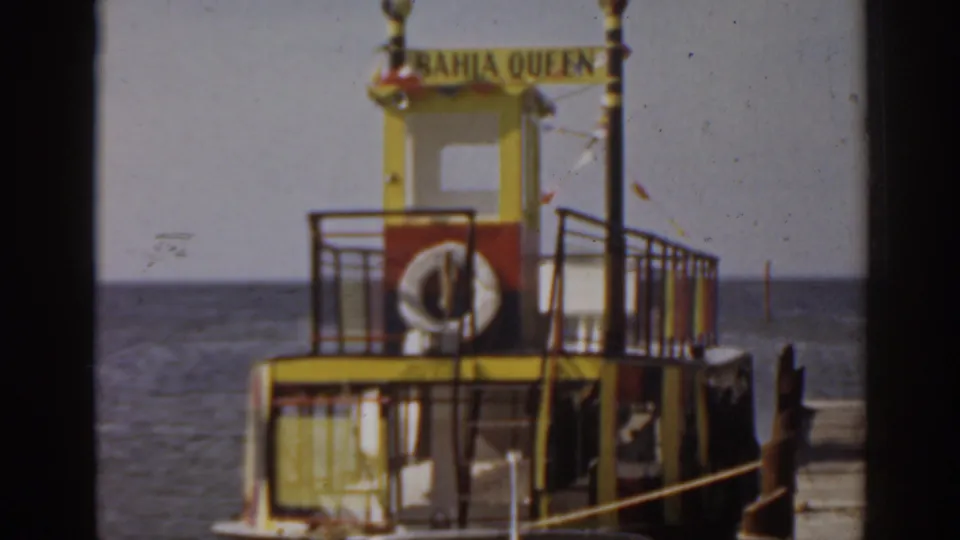 The Bahia Queen ferry appears docked at the water's edge, with yellow and red paint, railings, a life ring, and open sea behind it.